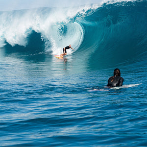 Two surfers in the ocean, one riding a wave.
